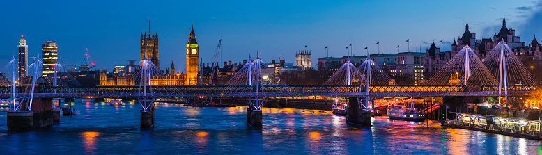 Panoramic view across the landmarks of central London, from the Southbank across the River Thames to the iconic towers of the Houses of Parliament and Big Ben illuminated against the blue dusk sky of the UK's vibrant capital city.