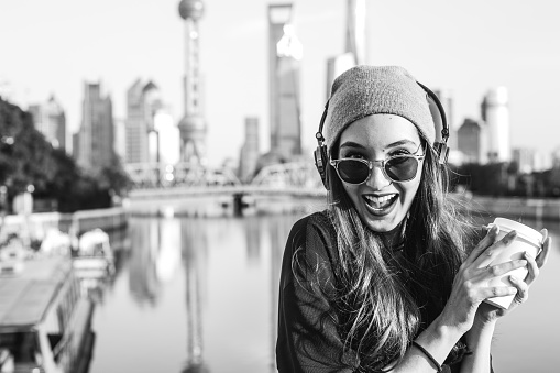 Beautiful young girl having fun in the city on a sunny spring day, enjoying the leisure time by the river with a coffee to go and music on her headphones.
