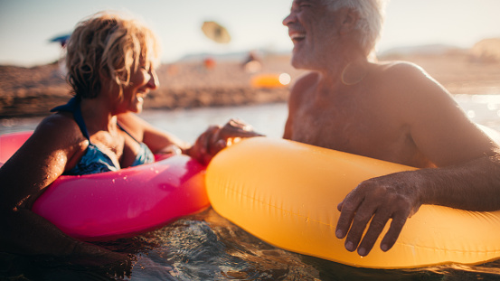 Photo of a cute, smiling senior couple being goofy while bathing in the sea with a swimming tubes. Wide photo dimensions.