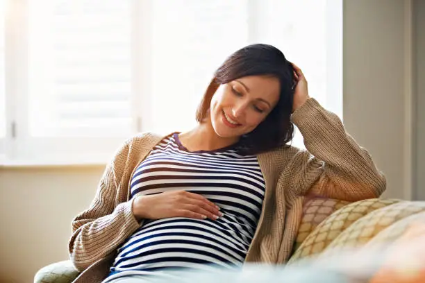 Shot of a smiling pregnant woman sitting on her sofa holding her stomach