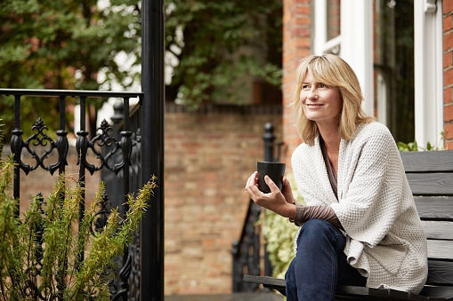 Shot of a mature woman sitting on a bench on her front porch drinking a coffee