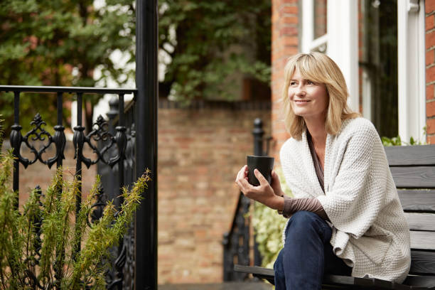 Relaxing into the day with a cup of coffee Shot of a mature woman sitting on a bench on her front porch drinking a coffee front porch stock pictures, royalty-free photos & images