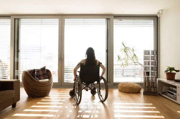 Beautiful young disabled woman in wheelchair at the window at home in her living room. Rear view.