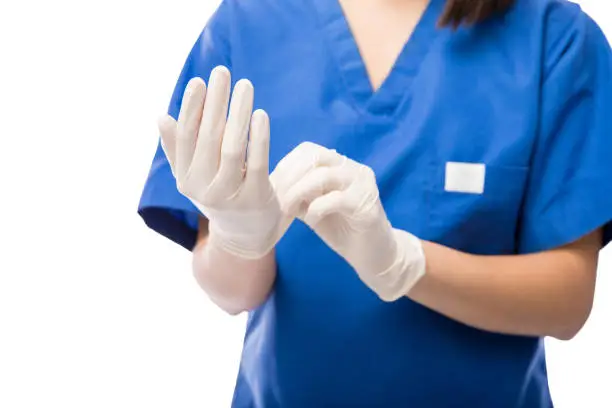 Closeup of a nurse in scrubs putting surgical gloves on while standing against a white background