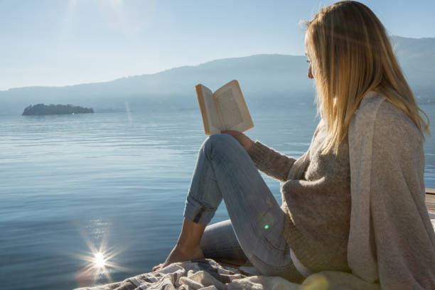 istock Young woman sits on jetty above lake, reading book 640241742