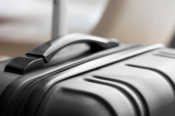 Close up of a modern, black, plastic suitcase with its handle extended above the case. It has a plastic carry handle and a textured finish. The case is standing beside a table. The selective focus is on the front left corner of the suitcase.