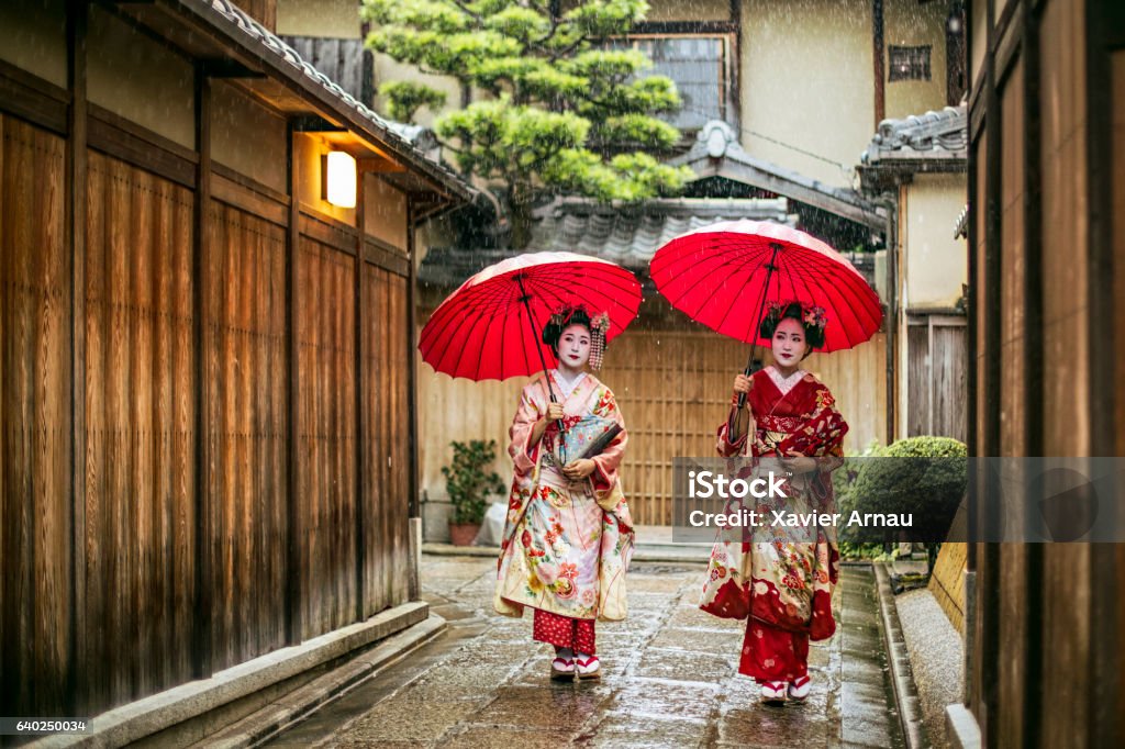 Geishas holding red umbrellas during rainy season Full length of young maikos holding red umbrellas during rainy season. Beautiful geisha girls wearing traditional dress called kimono. They are walking on wet street. Kyoto City Stock Photo