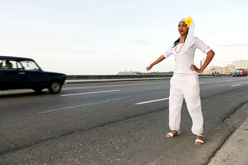 Beautiful, young Cuban woman calling taxi on the street in Havana, Cuba. In the background is visible the famous sea promenade El Malecon and an old car passing by. A woman wearing a Santeria white dress, La Habana Vieja, Havana, Cuba.