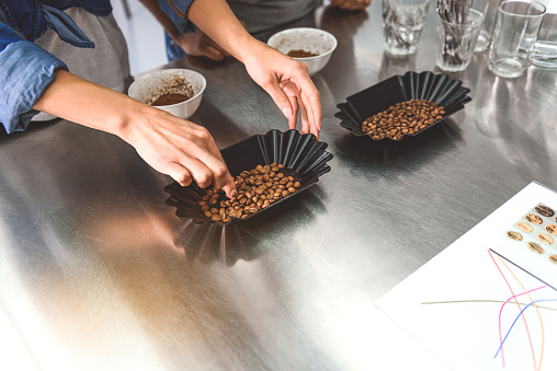 Black utensils with full beans are on table. Woman is taking few coffee seeds