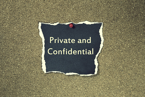 The phrase private and confidential on a black sticky note posted to a cork notice board