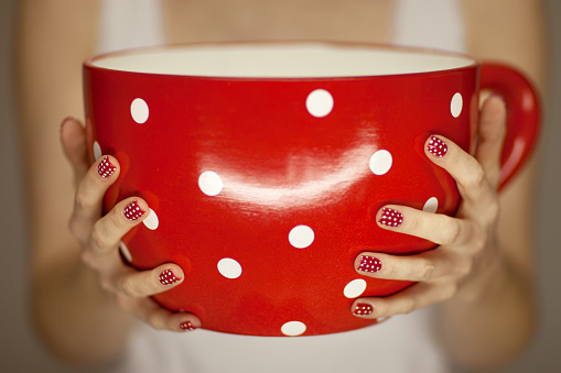 woman hands with red and white fingernails holding a big, giant  coffee cup in red with white dots.