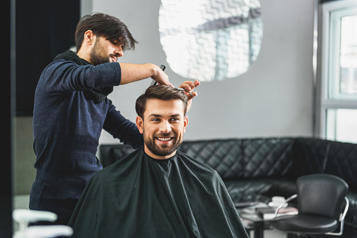 Professional stylist is serving customer at beauty salon. He is standing and cutting male hair with concentration. Young man is sitting and smiling