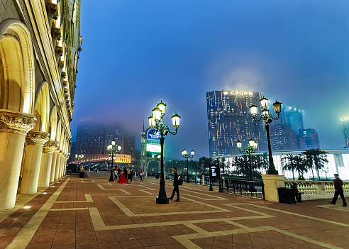 Macao, China - March 8, 2016: Embankment of Macau Casino and Hotel luxury resort in Macao in China. Late in the evening. Golden light illumination. City of Dreams and People are on the background