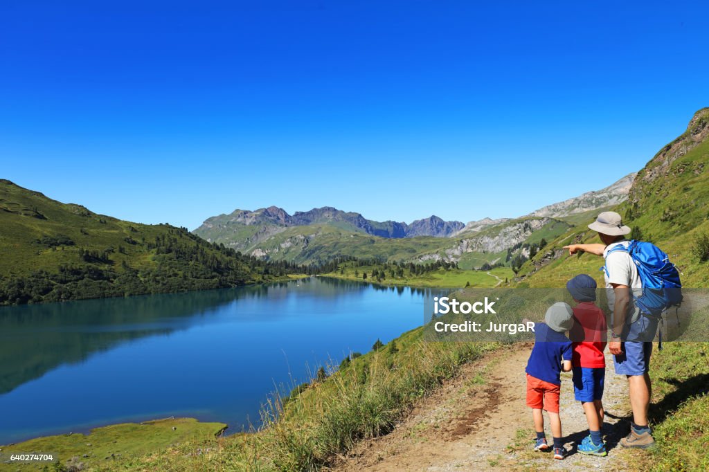 Family with Kids Hiking in the Swiss Mountains Father and his two sons are hiking in the mountains in Switzerland. The dad is wearing hiking clothes, a sun hat and is carrying a backpack. The boys are looking at a beautiful mountain lake, Engstlen Lake near Engelberg. Family Stock Photo