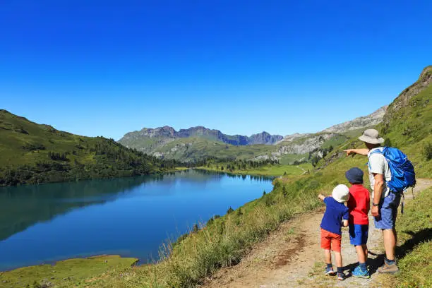 Father and his two sons are hiking in the mountains in Switzerland. The dad is wearing hiking clothes, a sun hat and is carrying a backpack. The boys are looking at a beautiful mountain lake, Engstlen Lake near Engelberg.