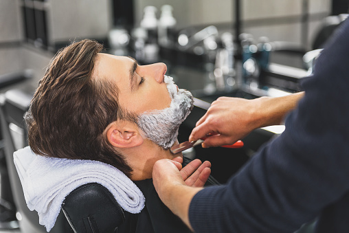 Young man is getting his stubble shaved by professional barber. He is sitting and relaxing