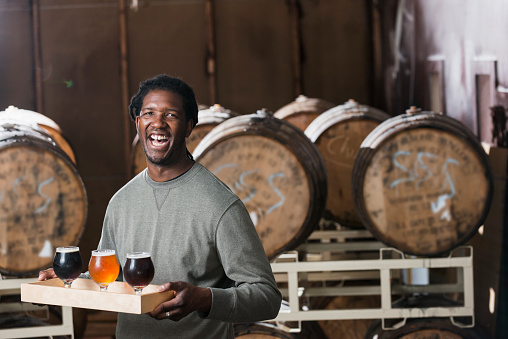 An African American man serving beer in a microbrewery.  He is smiling at the camera, carrying a tray with three glasses of craft beer.  Behind him are wooden barrels where the alcohol is stored.