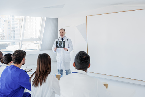 Senior doctor is teaching interns at clinic. He is standing and showing x-ray picture to them with friendly smile