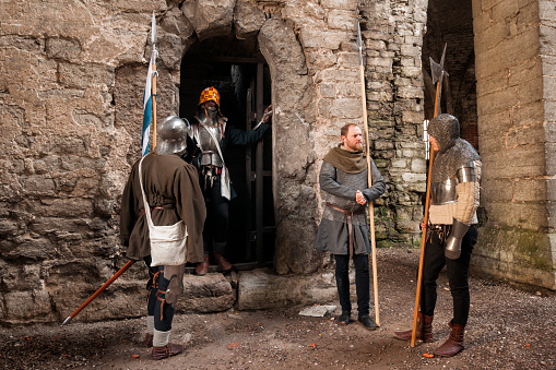 Four men wearing authentic medieval attire and weapons stand at the entrance of an ancient stone ruin that looks like a castle doorway, Visby, Gotland Island, Sweden, Europe