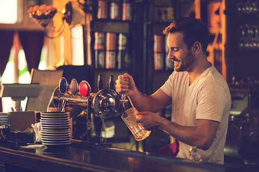 Young happy man working in a bar and pouring beer in a beer glass.