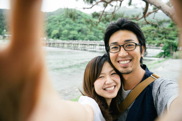 Cheerful Japanese couple taking selfie outdoors in a park Cheerful Japanese couple outdoors in a park taking selfie, bridge on the background. photographing herself stock pictures, royalty-free photos & images