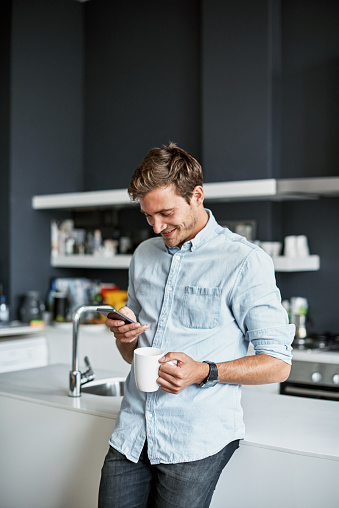 Shot of a young man using his cellphone and drinking coffee while relaxing at home