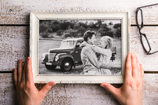 Hands of unrecognizable man looking at black-and-white photo of senior couple in white picture frame. Studio shot on white wooden background.