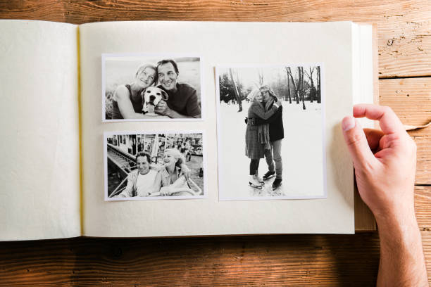 Hand holding photo album with pictures of senior couple. Studio Hand of unrecognizable person holding a photo album looking at various black and white pictures of senior couple. Studio shot on wooden background. photograph album stock pictures, royalty-free photos & images