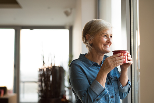 Beautiful senior woman at home standing at the window in her living room holding a cup of coffee or tea, smiling
