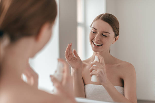 Body care. Over the shoulder view of young beautiful woman applying water spray while standing in front of the mirror semi dress stock pictures, royalty-free photos & images