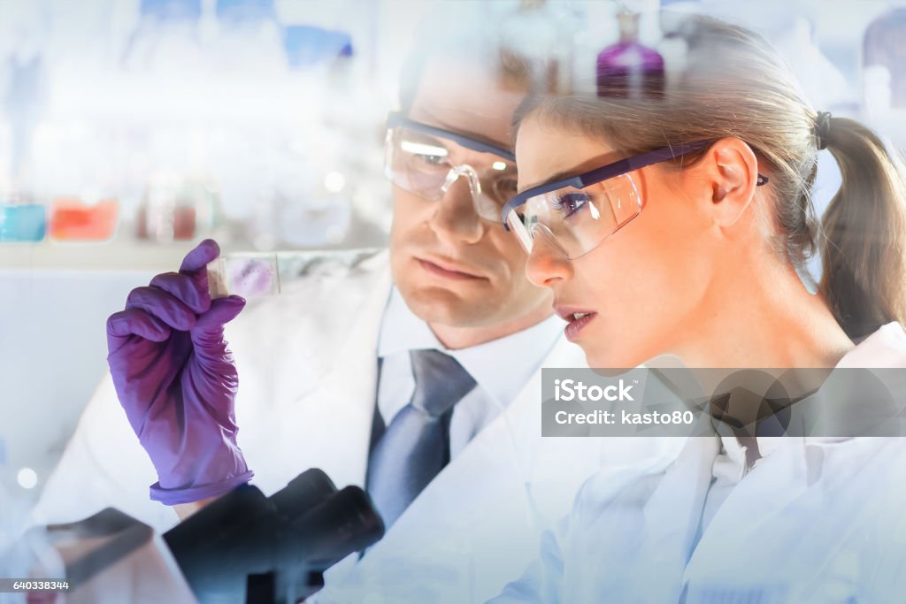 Health care professionals. Attractive young scientist and her post doctoral supervisor looking at the microscope slide in the forensic laboratory. Microscope Slide Stock Photo