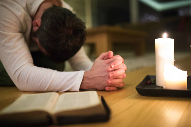 Young man praying, kneeling, Bible and candle next to him. Unrecognizable young man praying, kneeling on the floor, hands clasped together. Bible and burning candles next to him. Close up. kneeling stock pictures, royalty-free photos & images