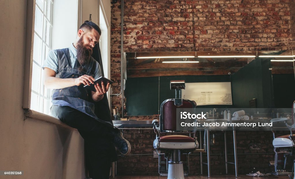 Hairstylist using digital tablet at his shop Hairstylist using digital tablet at his shop. Hairdresser sitting at barber shop working on digital tablet. Digital Tablet Stock Photo