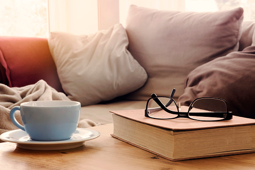 closeup of cup and book with eyeglasses on wooden table in front of couch in living room