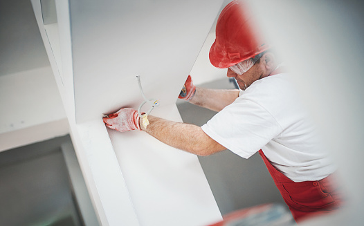 Closeup side view of a construction worker sanding a drywall and preparing it for painting. He's wearing a red uniform and a red helmet, also has protective glasses.