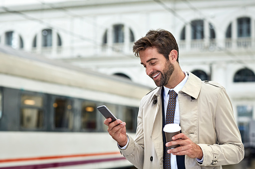 Smiling handsome businessman using smart phone. Male professional is holding coffee cup. He is standing at railroad station.
