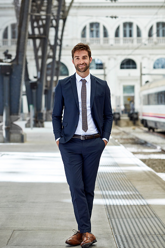 Full length of businessman standing with hands in pockets. Confident professional is wearing suit. He is standing at railroad station platform.