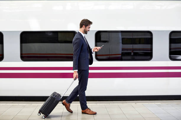Side view of businessman using phone at station Side view of businessman using phone at station. Full length of male is pulling wheeled luggage by train. He is wearing suit. railroad station platform stock pictures, royalty-free photos & images