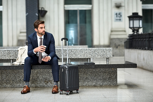 Thoughtful businessman having coffee while holding phone. Male is sitting by wheeled luggage on concrete bench. He is in formalwear.