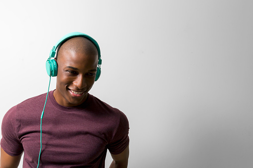 Smiling man listening music through headphones. Young male is standing against white background. He is wearing maroon t-shirt.