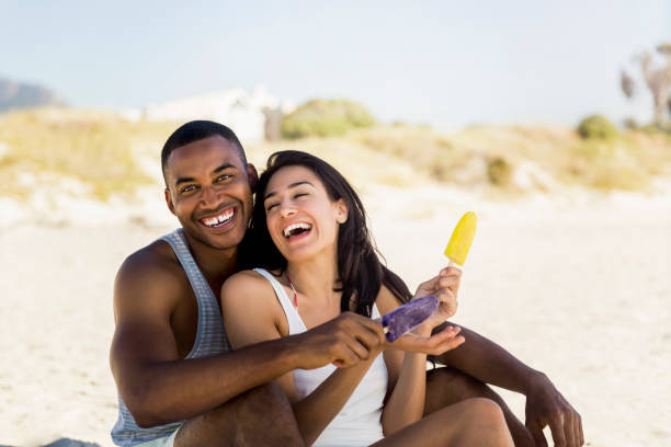 Cheerful couple holding popsicles while sitting at beach. Portrait of happy man sitting with woman on sunny day. They are enjoying summer vacation.