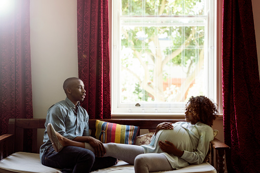 A photo of man talking to pregnant woman at brightly lit home. Couple is sitting on sofa by window. They are in casuals.
