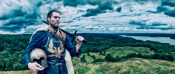 Warrior with helmet and sword stands outside overlooking fields and a big lake. His sword rests on his shoulder and he looks serious.