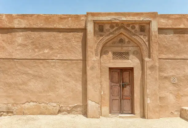 A traditional arched doorway with gypsum carvings and a carved door stands in bright sunlight in a wall of an old fort in the Arabian Gulf.