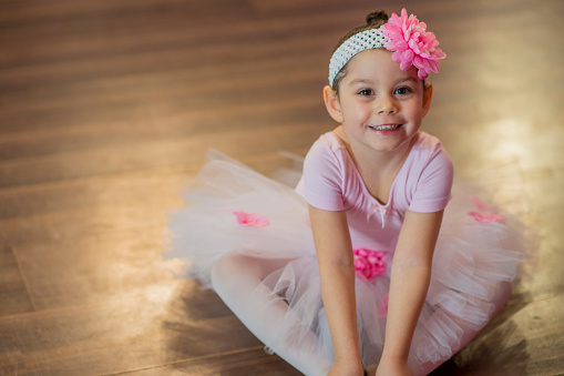 A cute little girl is in ballet class and is wearing fake flowers and a tutu. She is smiling and looking at the camera.