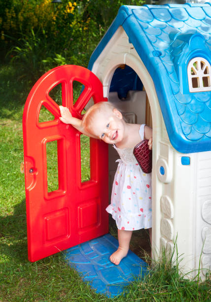 Toddler in kids playhouse outdoor Little baby girl wearing white dress looking out from plastic play house doorway in a summer playground kids play house stock pictures, royalty-free photos & images