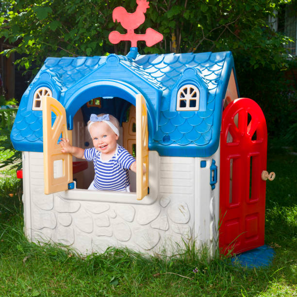 Toddler in kids playhouse outdoor Little baby girl wearing white-blue striped summer dress looking out from plastic play house window in a playground playhouse stock pictures, royalty-free photos & images