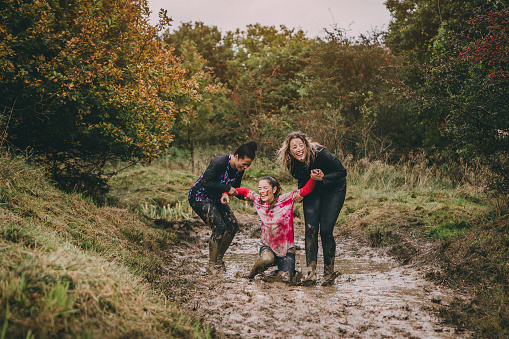 Women are taking part in a charity obstacle course. One woman is stuck in a muddy ditch and the other two are trying to help her get out. They are laughing to hard to be able to lift her.