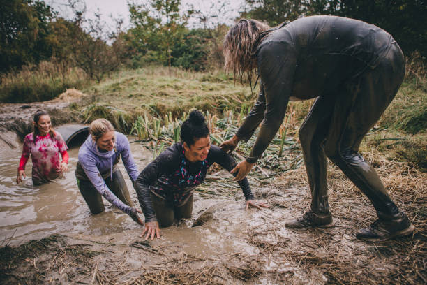 Obstacle Course Support Women are taking part in a charity obstacle course. They are wading through a muddy ditch and one of the women is standing and helping them get out. obstacle course stock pictures, royalty-free photos & images