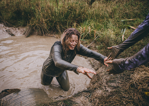 Woman is being pulled out of a ditch full of muddy water by her friend. They are both taking part in a charity obstacle course.